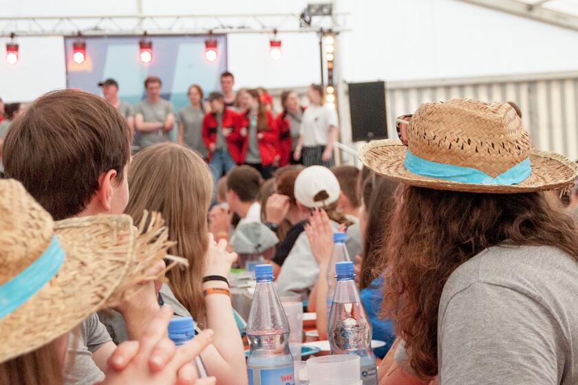 Jugendliche sitzen an einem Tisch in einem großen Zelt und schauen nach vorne auf eine Bühne.