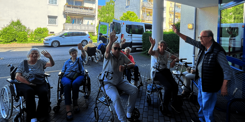Menschen stehen mit Rollatoren unter einem Vordach und winken