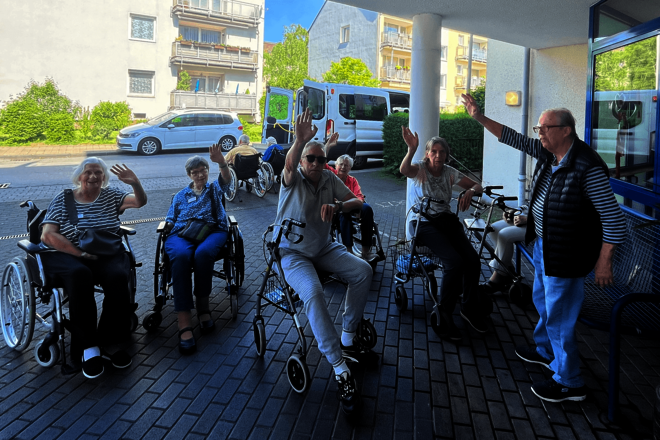 Menschen stehen mit Rollatoren unter einem Vordach und winken