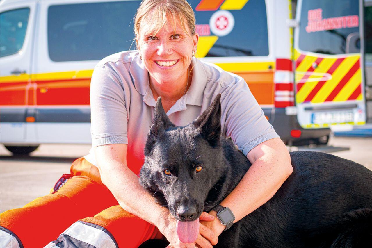 Johanniter-Mitarbeiterin in grauem Poloshirt kniet vor Einsatzfahrzeug und hält einen schwarzen Hund.