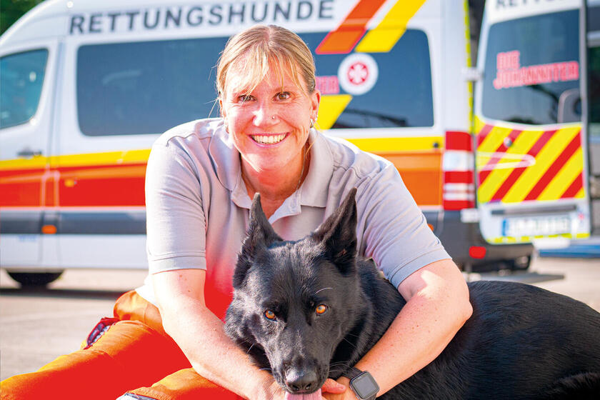 Eine Frau sitzt auf dem Boden und umarmt einen Hund vor einem Johanniter-Einsatzfahrzeug mit der Aufschrift „Rettungshunde“.