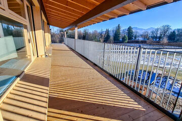 Langer, überdachter Balkon aus Holz mit großen Fenstern und Bergpanorama im Hintergrund.