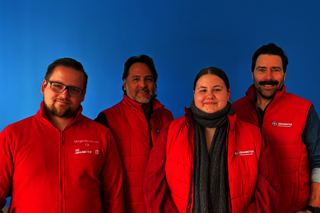 Vier Mitarbeitende des Johanniter Mitgliederservice in roter Einsatzkleidung stehen vor einer blauen Wand.