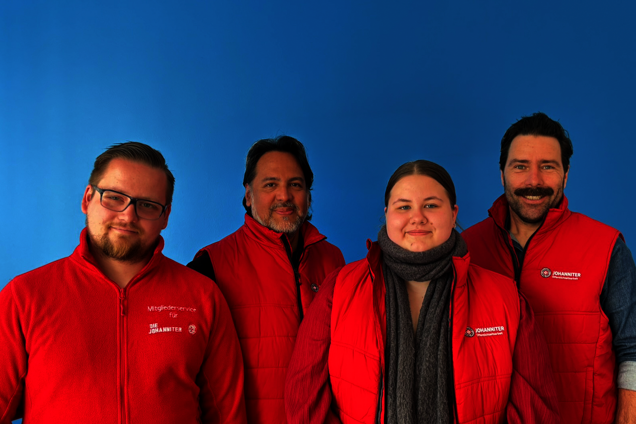 Vier Mitarbeitende des Johanniter Mitgliederservice in roter Einsatzkleidung stehen vor einer blauen Wand.