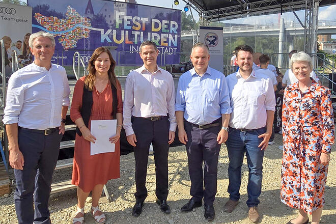 Mehrere Leute stehen vor einer Bühne. Auf der Bühne ist ein großes Plakat mit der Aufschrift „Fest der Kulturen Ingolstadt“.