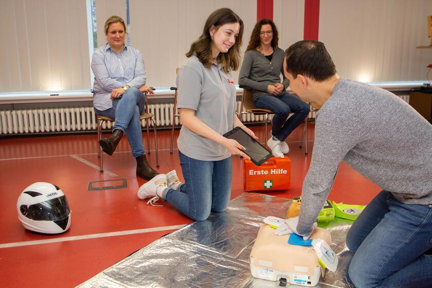 Zu sehen sind vier Personen. Zwei Frauen sitzen im Hintergrund auf Stühlen und schauen in die Mitte. Dort stützt ein Mann seine Arme auf den Herzbereich eines Puppentorsos, während eine Erste-Hilfe-Ausbilderin ihm einen Laptop zum Draufschauen hinhält.