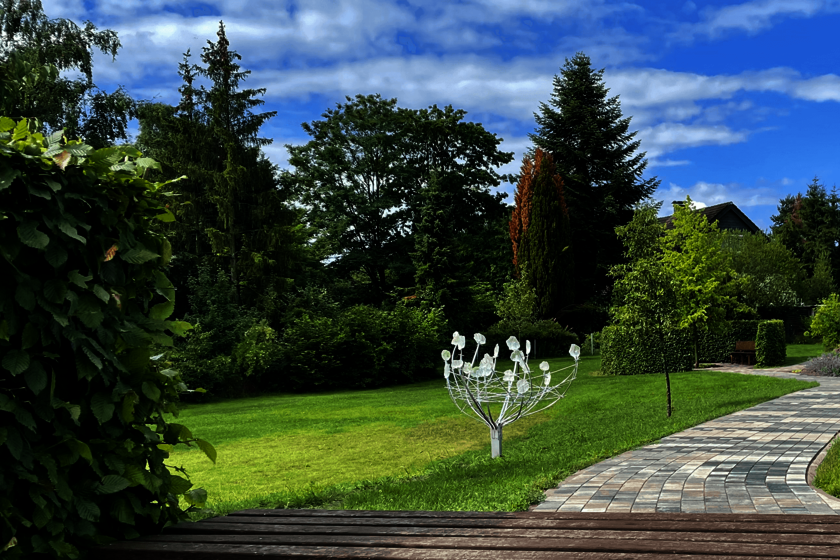 Im Hintergrund Bäume und Sträucher, im Vordergrund eine hözerne Sitzfläch eeine Bank. Im Mittelpunkt des Bildes ist eine gläserne Skulptur in der Form eines Straußes zu sehen.