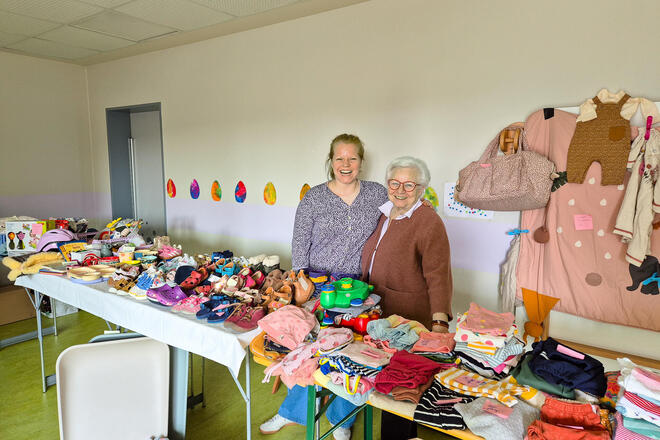 In einem Raum stehen eine Frau und eine ältere Frau hinter einem Verkaufsstand. Dieser ist voll mit Bekleidung.