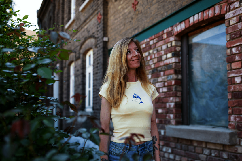 Frau steht vor einem Backsteingebäude und blickt nach oben, umgeben von Pflanzen.  