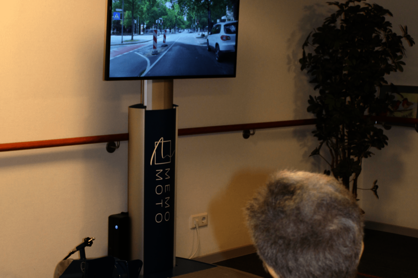 Eine Person ist von hinten dabei zu sehen, wie sie auf einen Bildschirm blickt, auf dem eine Straße zu sehen ist. Links unten im Bild sieht man ein Fußfahrrad.