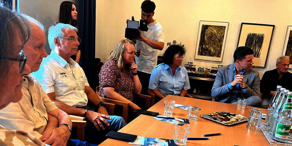 mehrere Menschen sitzen rundherum an einem Tisch. Darauf liegen Handys.