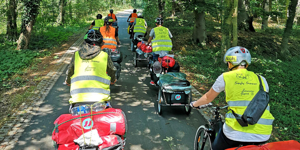 Mehrere Jugendliche auf Fahrrädern mit Helm, Warnweste und Gepäcktaschen. Sie fahren durch ein Waldstück.