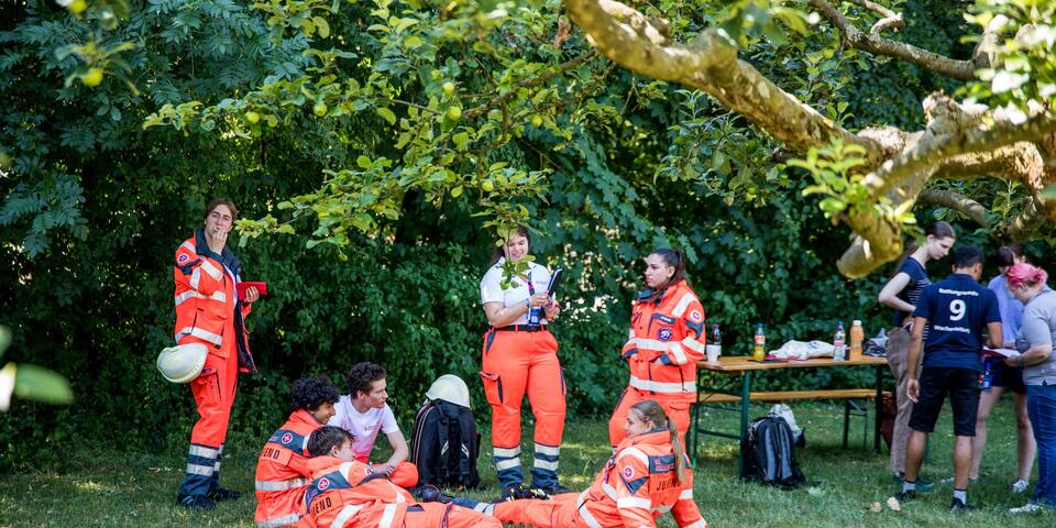 Mehrere Einsatzkräfte und Helfer*innen in orangefarbener Einsatzkleidung ruhen, sprechen und bereiten sich im schattigen Park auf Einsätze oder Übungen vor.