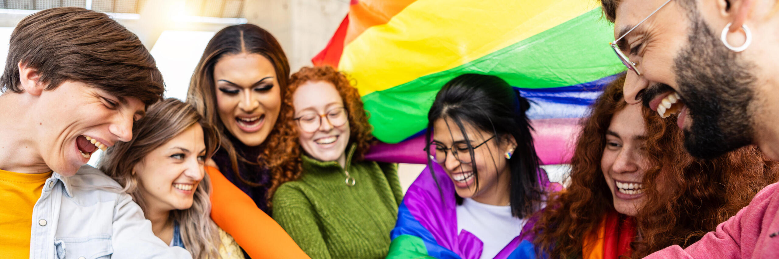 Junge Menschen verschiedenster Identitäten schlagen zusammen im Kreis ein, im Hintergrund weht eine Regenbogenflagge.