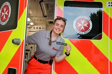 Junge Johanniterin lächelt in der Tür eines Rettungswagens und hält ein Namensschild