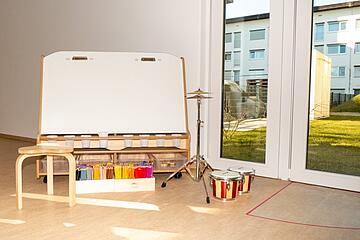 Weiße Leinwand auf einem Holzgestell. Davor steht ein Holzstuhl und eine Box mit buten Stiften. Rechts daneben stehen zwei kleine Trommeln sowie ein Schlagzeug-Becken.