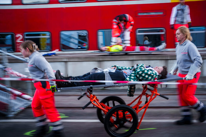 Zwei Johanniter-Rettungskräfte transportieren eine liegende Person auf einer Trage entlang eines Bahnsteigs; Absperrband im Vordergrund und ein stehender Zug im Hintergrund, weitere Verletzte werden versorgt.