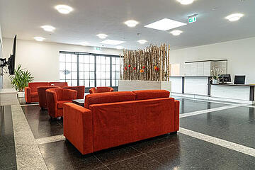 Moderner Aufenthaltsraum mit roten Sofas, Pflanzen und Empfangstisch im Hintergrund.