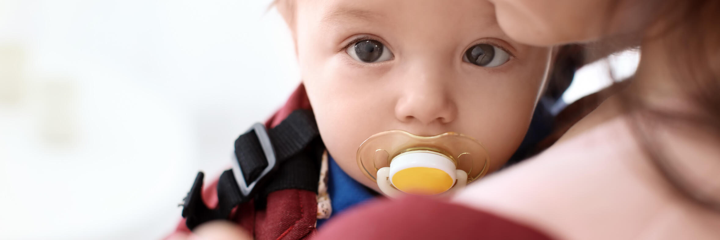 Baby mit Schnuller schaut über die Schulter einer Frau, die es im Tragetuch hält.