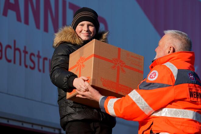 Ein Kind, das fröhlich lächelnd vor einem Johanniter-LKW steht, erhält ein Weihnachtstrucker-Paket von einem Johanniter