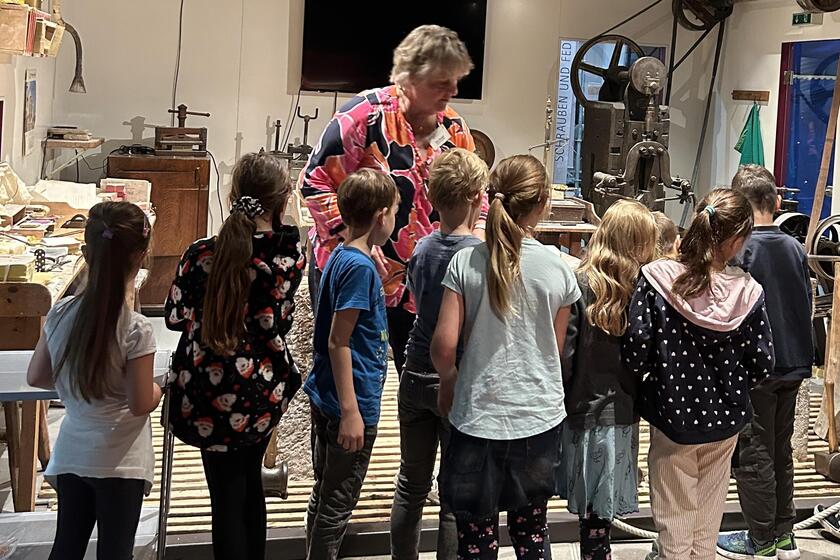 In der Goldwerkstatt stehen neun Grundschulkinder vor einer Museumsleiterin. Im Hintergrund ist altes Werkzeug zu erkennen und ein Bildschirm.