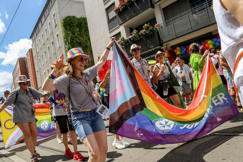 Johanniter nehmen an einer Pride-Parade teil und halten eine große Johanniter-Pride-Flagge. Viele tragen graue Johanniter Polos und bunte Hüte. Im Hintergrund feiert die Menge vor einem Haus mit dem Schild „Queeres Zentrum“.