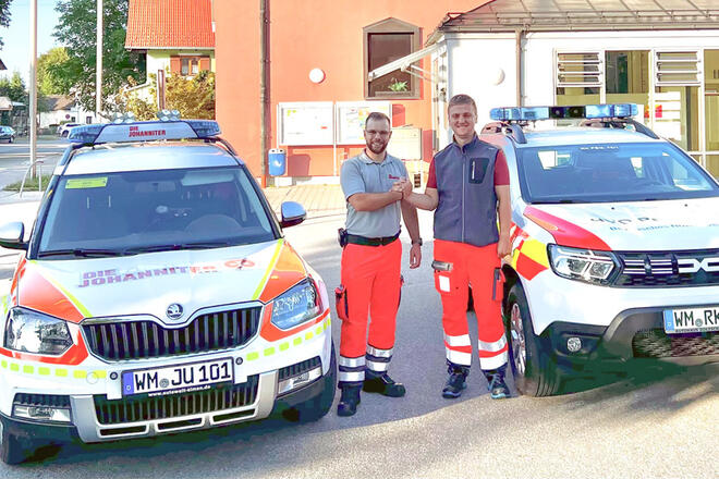 Ein Johanniter und ein Rotes Kreuz Mitarbeiter schütteln Hände und stehen zwischen ihren beiden Einsatzfahrzeugen.