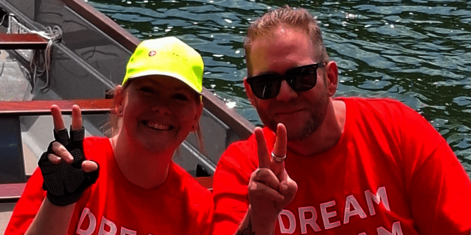 Zwei Personen in roten T‑Shirts zeigen Victory‑Zeichen und lächeln im Boot.