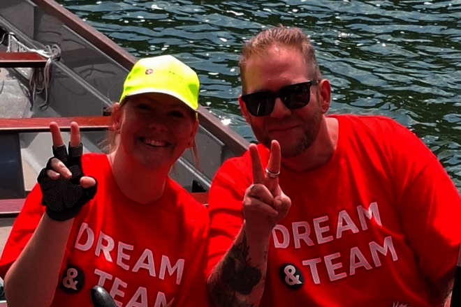 Zwei Personen in roten T‑Shirts zeigen Victory‑Zeichen und lächeln im Boot.