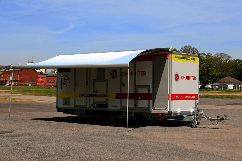 Ein großer Fahrzeuganhänger mit rot-gelben Streifen und einem Vordach steht frei auf einem großen Platz. 