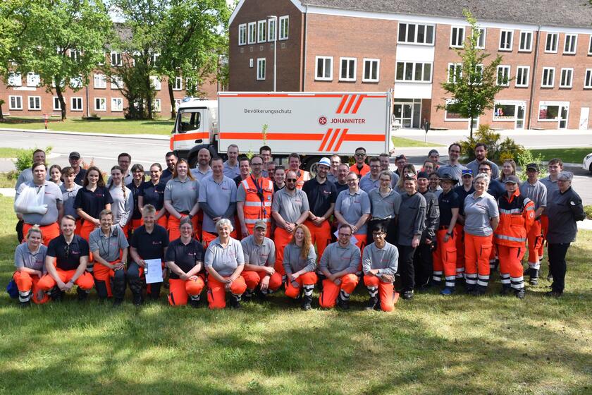 65 ehrenamtlich Helfende Johanniter sitzen in großer Gruppe auf einer Wiese zusammen