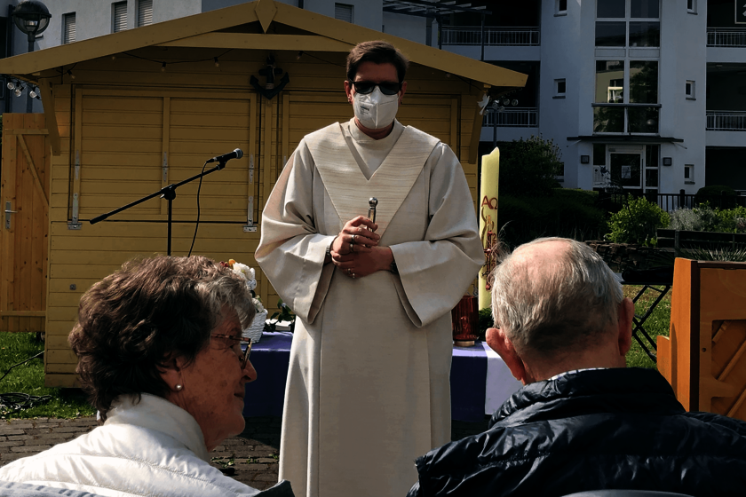 Ein älteres Paar sitzt im Freien einem weißgekleideten Geistlichen gegenüber.