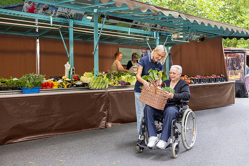 Eine Johanniterin reicht einer Frau, die in einem Rollstuhl sitzt, einen Korb gefüllt mit Gemüse. Sie befinden sich vor einem Marktstand.