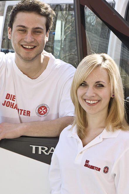Junge Frau und junger Mann in weißen T‑Shirts mit Johanniter‑Logo stehen lächelnd an der offenen Fahrertür eines Einsatzfahrzeugs.