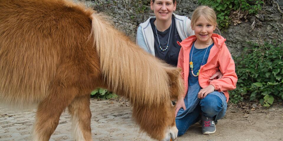 Mutter und Tochter sitzen bei Pony welches gerade aus einer Schale frisst