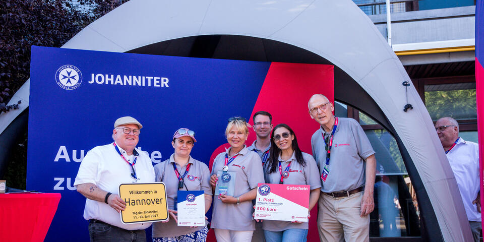 Gruppe von Johannitern und Gästen steht vor einer Messewand und präsentiert Urkunden, einen Gutschein und einen Scheck für den ersten Platz.