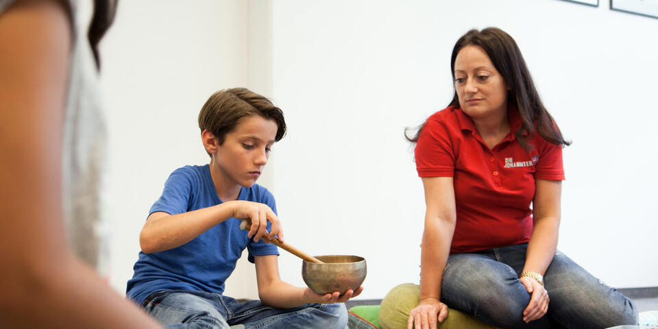 Ein Junge schlägt eine Klangschale, neben ihm eine Trauerbegleiterin, die ihn aufmerksam beobachtet.