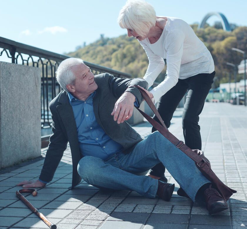 Mann ist gestürzt und liegt am Boden. Frau packt seinen Arm und versucht ihm aufzuhelfen.