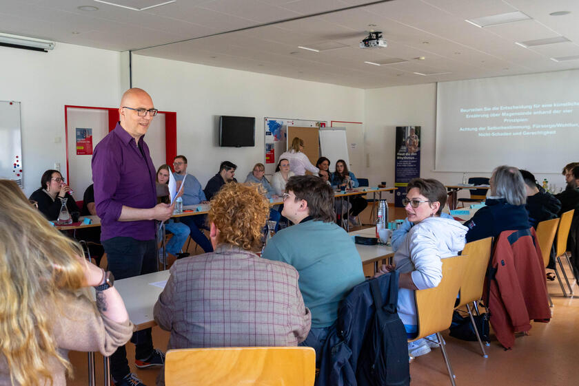 Der Referent Sebastian Heinlein verteilt Zettel an die sitzenden Workshopteilnehmenden.