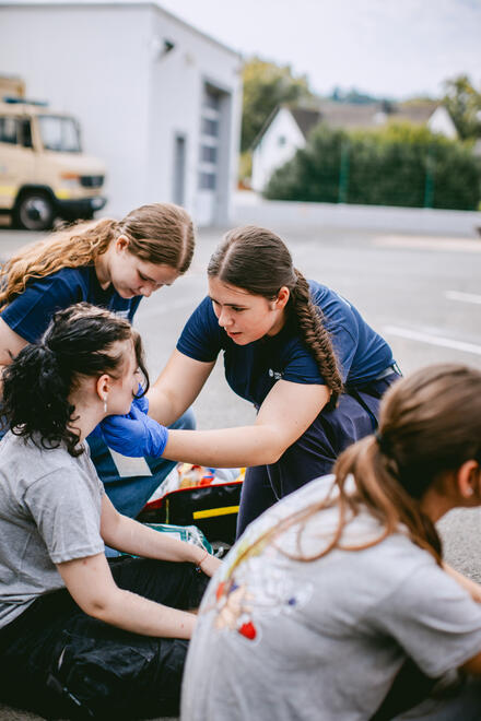 Zwei Schulsanitäterinnen versorgen die Wunde einer Mitschülerin auf dem Pausenhof.