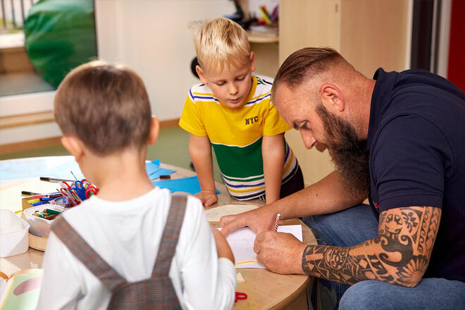 Ein Mann mit langem Bart und Tattoos am Arm sitzt mit 2 kleinen Jungen an einem Basteltisch. Der Mann zeichnet etwas, während die Kinder ihm neugierig zusehen.