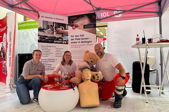 Ein Mann in Einsatzkleidung und zwei Frauen mit grauem Johanniter-Polo knien an einem Messestand