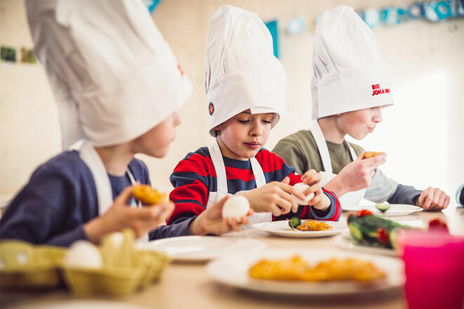 3 Kinder mit Johanniter Kochschürzen- und Mützen sitzen an einem Tisch und essen.