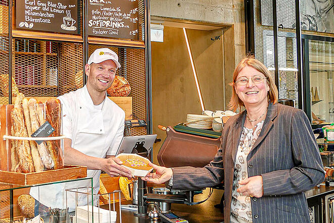 Einer Frau im grauen Blazer wird gerade von einem Bäcker in weißer Arbeitsuniform ein Gebäck überreicht. Sie stehen in einer Bäckerei, er hinter dem Tresen und beide lächeln in eine Richtung.