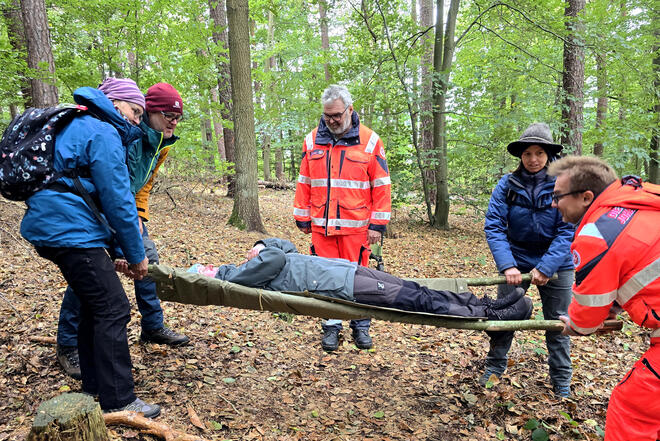 Die Teilnehmer eines Outdoor-Erste-Hilfe-Kurses transportieren einen der Kursteilnehmer mit einer aus Stöcken gefertigten Trage.