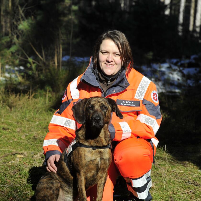 Eine Johanniter-Rettungshundeführerin kniet in auffälliger Einsatzkleidung im Wald und lächelt in die Kamera. Neben ihr sitzt ihr braun-schwarzer Rettungshund, beide wirken aufmerksam und einsatzbereit.