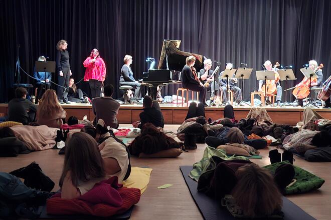 Eine kleinere Orchesterbesetzung spielt auf einer Bühne vor einem liegenden Publikum, das auf Matten und Decken entspannt zuhört.