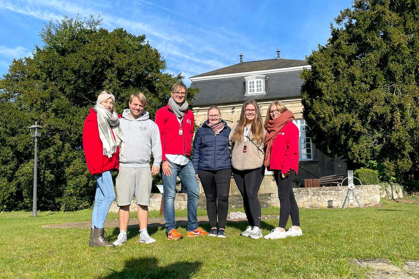 Sechs motivierte junge Erwachsene in Johanniter-Jugend-Kleidung stehen vor einem Landhaus auf einer Wiese und blicken in die Kamera.