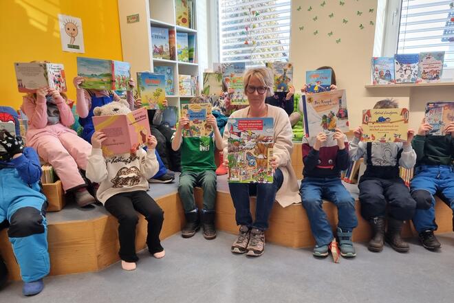 Vorschulkinder sitzen in der Bücherei und halten Bilderbücher vor ihr Gesicht; eine erwachsene Frau sitzt in der Mitte und zeigt das Johanniter‑Wimmelbuch.