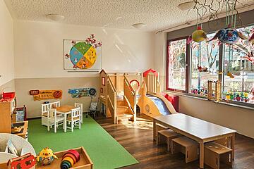 Heller Gruppenraum mit Spielbereich, kleinen Tischen, Holz-Kletterelementen und großen Fenstern.